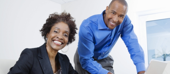 A smiling woman in a blazer and a man in a blue shirt working at a laptop