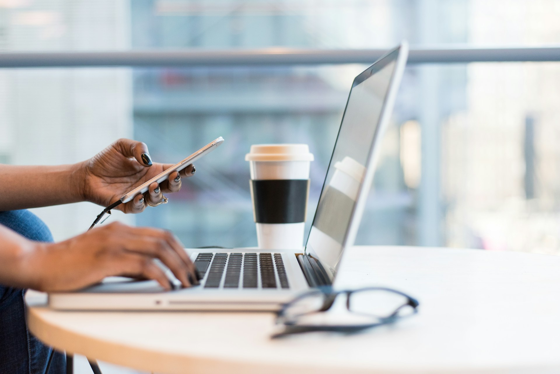 Hands on a laptop and a phone beside a coffee cup and glasses