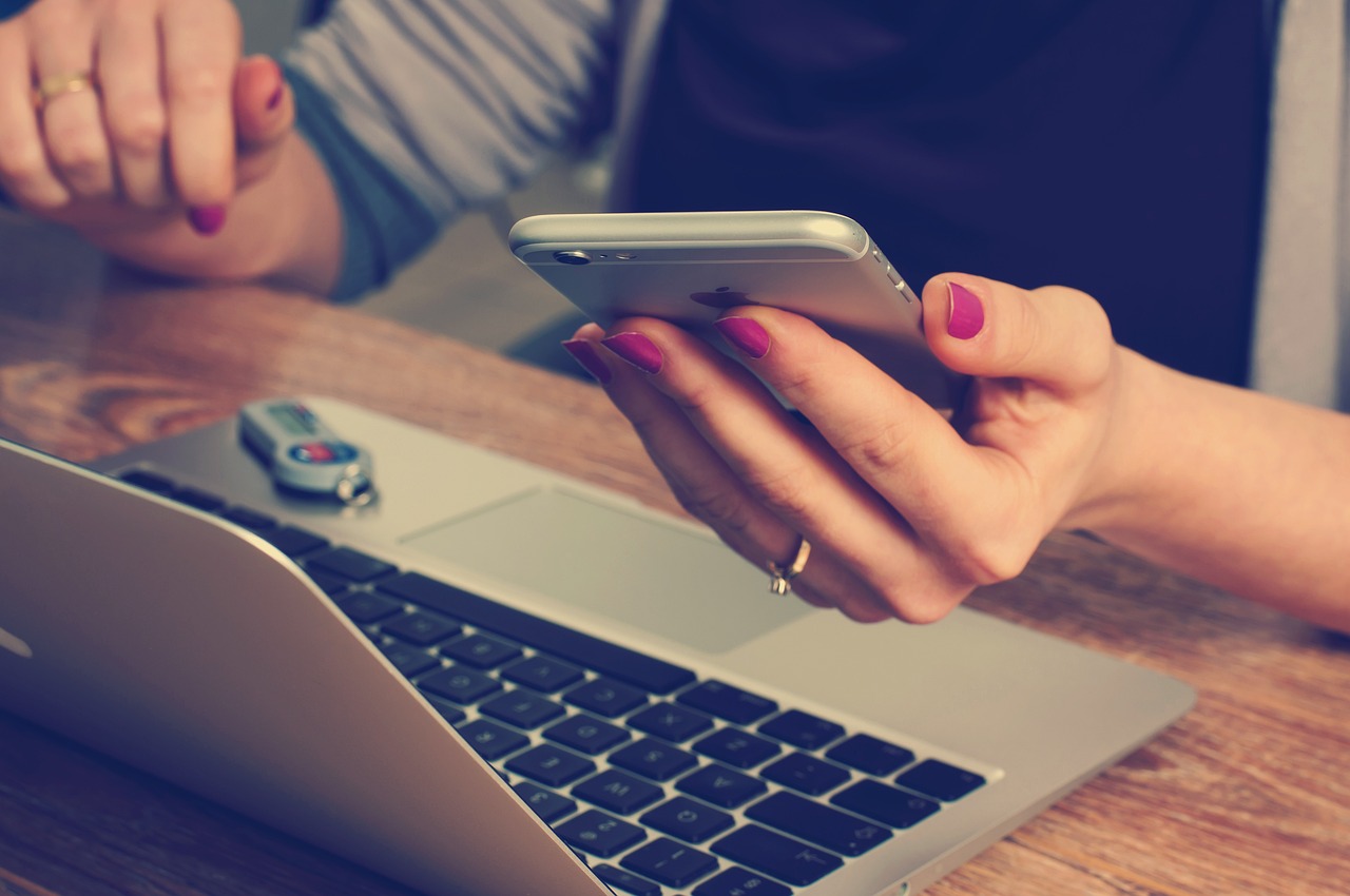Hands holding a smartphone above an open laptop on a wooden desk