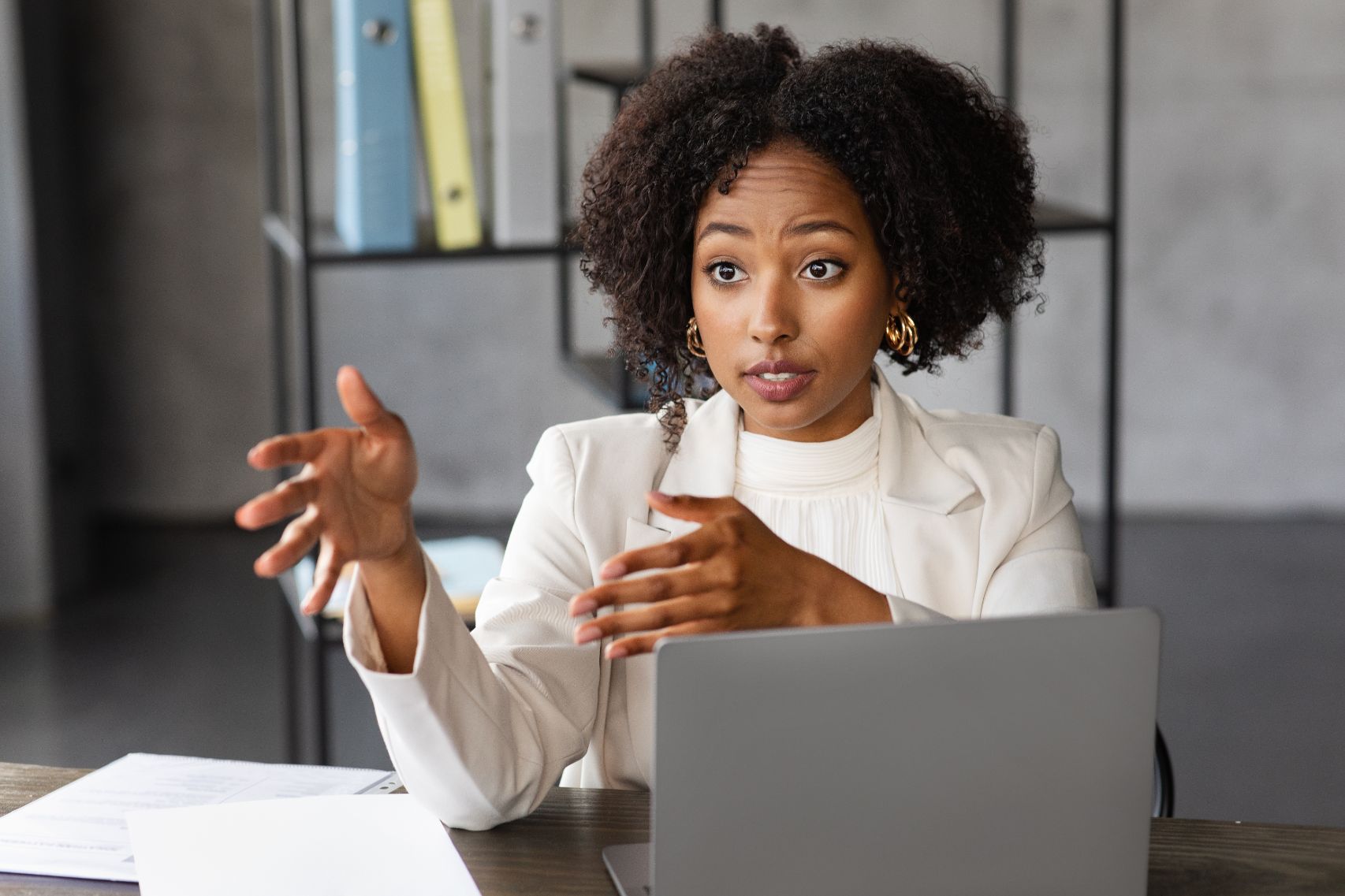A woman in a white blazer talking and gesturing at a laptop
